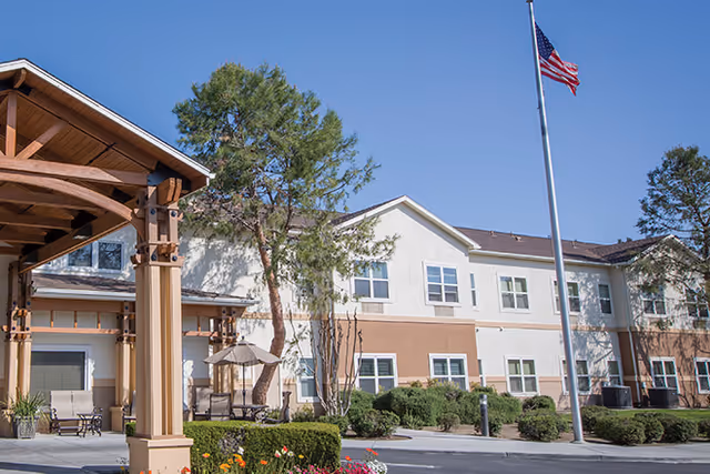 Exterior view of a two-story senior living facility building with beige and white walls, multiple windows, and a covered entrance supported by wooden beams. There is a flagpole with an American flag in front of the building, surrounded by landscaped bushes, trees, and flowers under a clear blue sky.