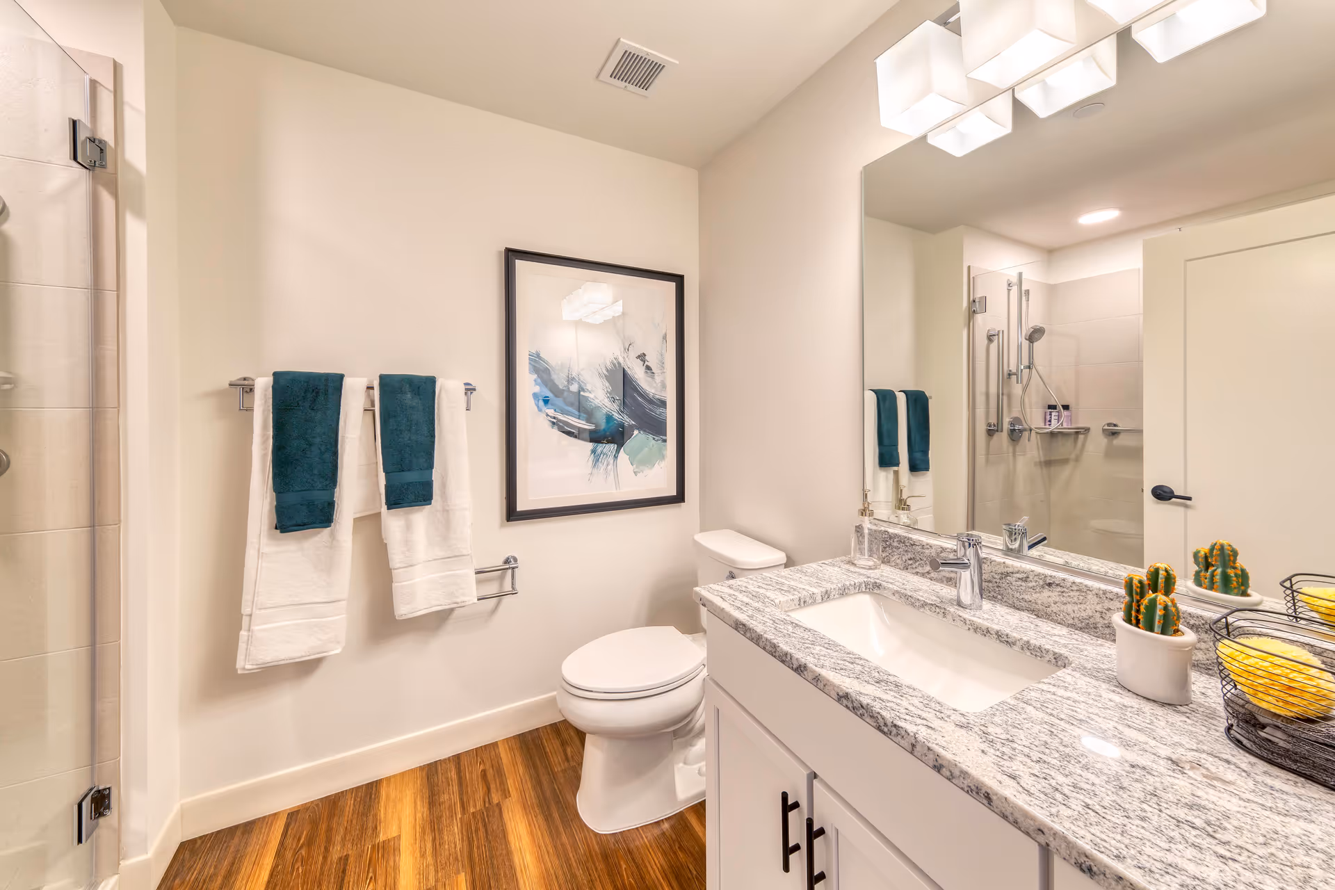 Modern bathroom with a white toilet, granite countertop with an under-mount sink, and a large mirror above the sink. Two white towels with dark teal hand towels hang on a towel rack on the wall. A framed abstract artwork is mounted above the toilet. The floor is wood, and a glass shower door is partially visible on the left side. A small potted cactus and a wire basket with yellow sponges sit on the countertop.