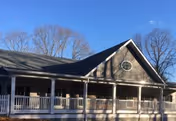 Exterior view of a single-story building with a covered porch and white railing, set against a clear blue sky with leafless trees in the background.