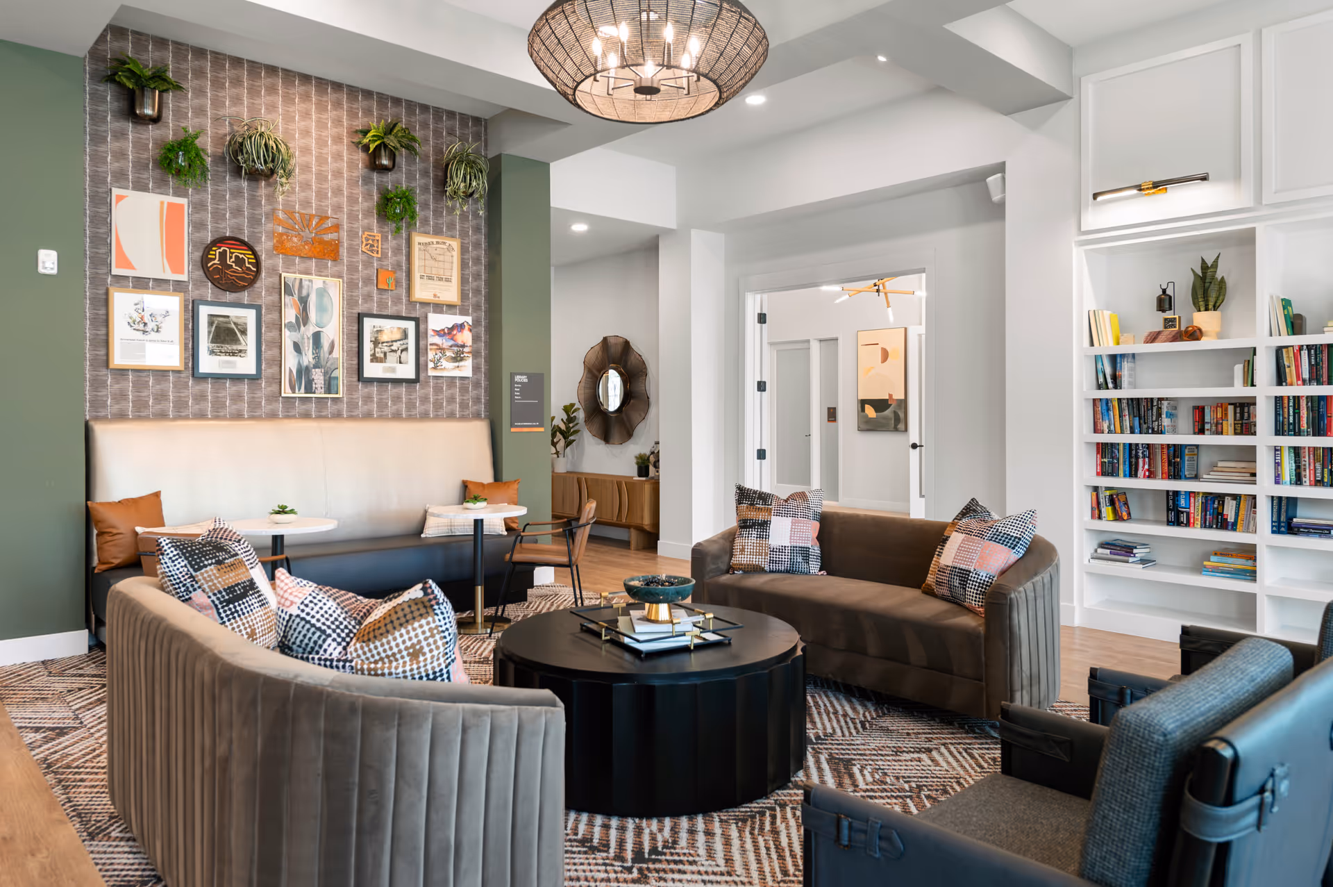 A modern and cozy living room area with two brown velvet sofas adorned with patterned pillows, a round black coffee table with decorative items, and a patterned rug. The back wall features a green accent wall with various framed artworks and hanging plants. There is a built-in white bookshelf filled with books on the right side and a doorway leading to another room in the background.