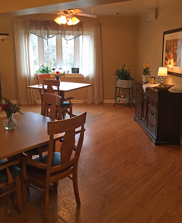 A cozy dining room with wooden floors and beige walls. There are two wooden tables with chairs around them, each table decorated with small vases of flowers. A large window with sheer curtains lets in natural light. On the right side, there is a dark wooden sideboard with a lamp, flowers, and decorative items. A ceiling fan with lights is mounted above.