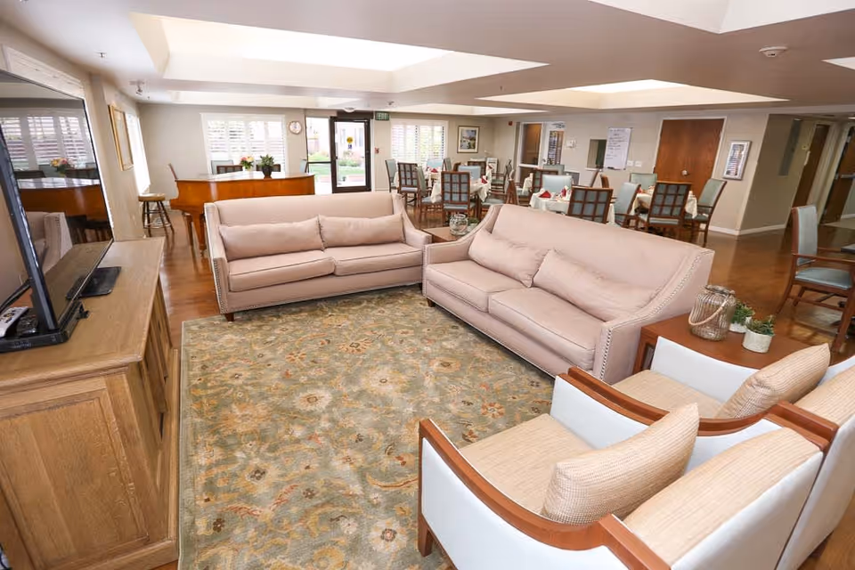 A spacious senior living common area featuring two beige sofas, two armchairs, a patterned area rug, a wooden TV stand with a flat-screen TV, and a grand piano near large windows with white shutters. In the background, there are several dining tables with chairs and place settings, and a door leading outside.