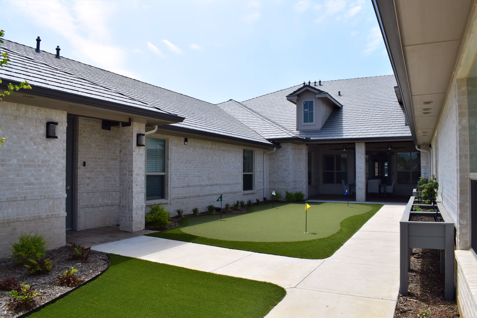 Outdoor courtyard area of a senior living facility with a small artificial putting green featuring several colored flags, surrounded by light-colored brick buildings with windows and a covered patio area with chairs and tables.