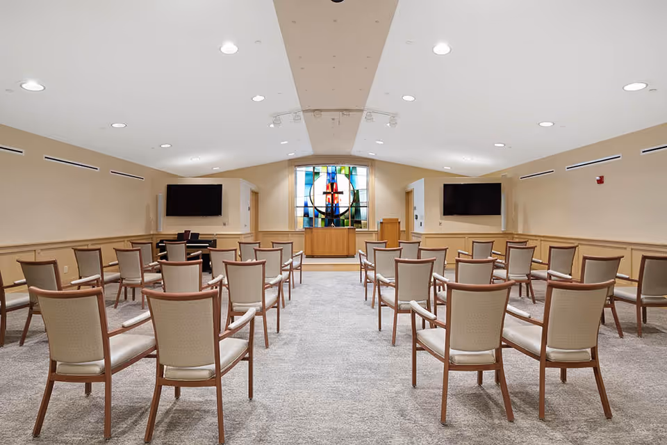 A spacious room with rows of beige cushioned chairs arranged facing a podium and a colorful stained glass window at the front. Two large flat-screen TVs are mounted on the side walls. The room has a neutral color scheme with beige walls and carpeted flooring, and recessed lighting in the ceiling.