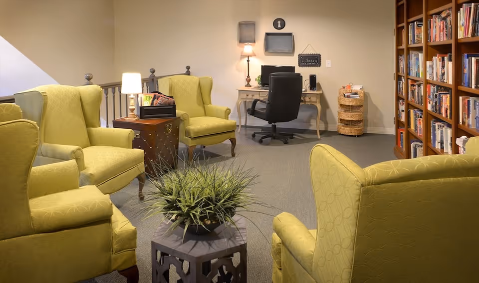A cozy sitting area with four green upholstered armchairs arranged around a small table with a potted plant. In the background, there is a desk with a black office chair, a lamp, and a computer. To the right, there is a tall bookshelf filled with books and a wicker basket. The walls are painted beige, and the floor is carpeted.