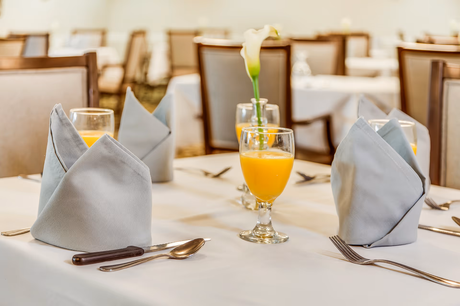 A dining table set with folded gray napkins, glasses of orange juice, silverware, and a single white flower in a vase in the center. The background shows more tables and chairs in a softly lit dining room.
