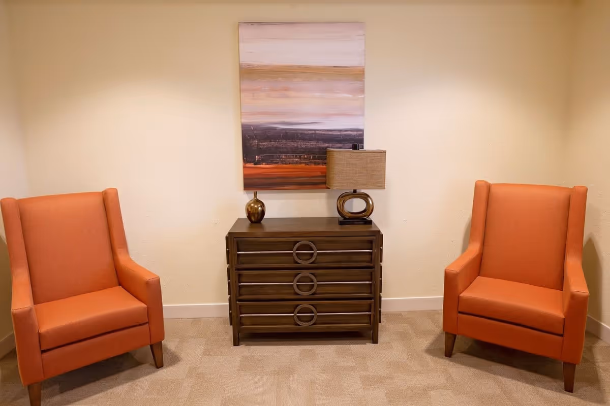 A small seating area with two orange upholstered armchairs facing forward, separated by a dark wooden chest of drawers with three circular handles. On top of the chest is a modern table lamp with a beige shade and a small decorative vase. Above the chest hangs an abstract painting with horizontal bands of muted colors including beige, brown, and orange. The walls are light beige and the floor is carpeted in a neutral tone.
