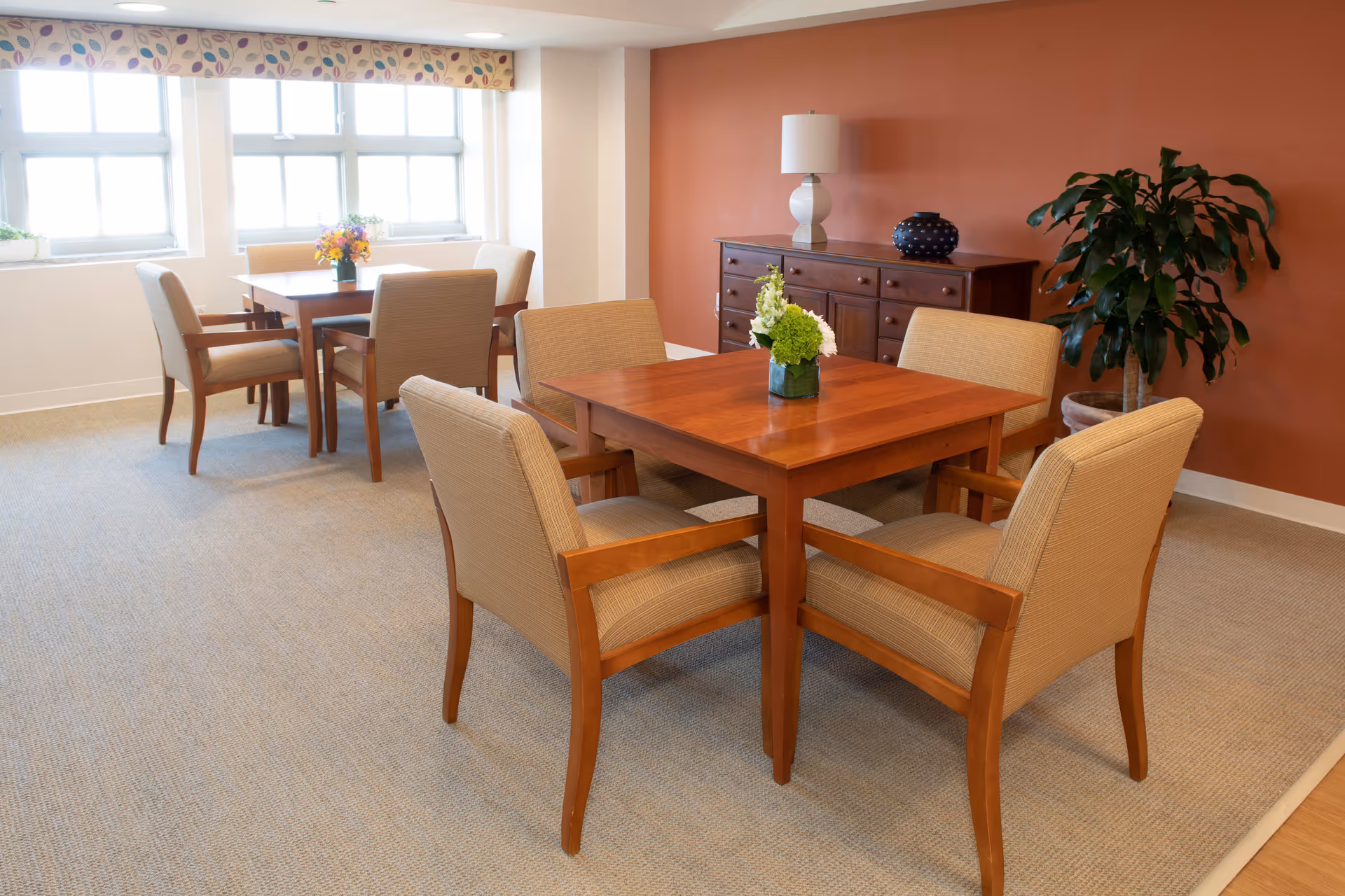 A bright dining room with two wooden tables, each surrounded by four beige upholstered chairs. The room has large windows with a floral valance, a wooden sideboard with a lamp and decorative vase, and a large potted plant against a warm orange wall. Each table has a small floral centerpiece.