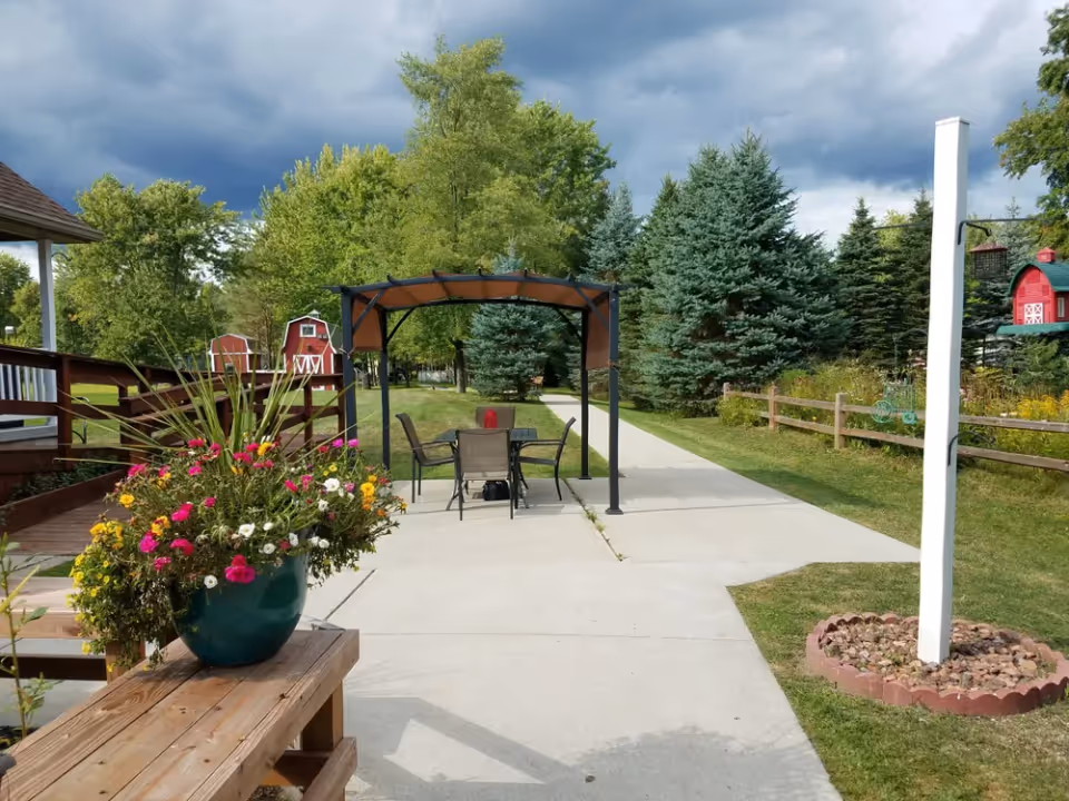 Outdoor patio area with a covered table and chairs, a large potted flower arrangement, walkway and trees under a cloudy sky.