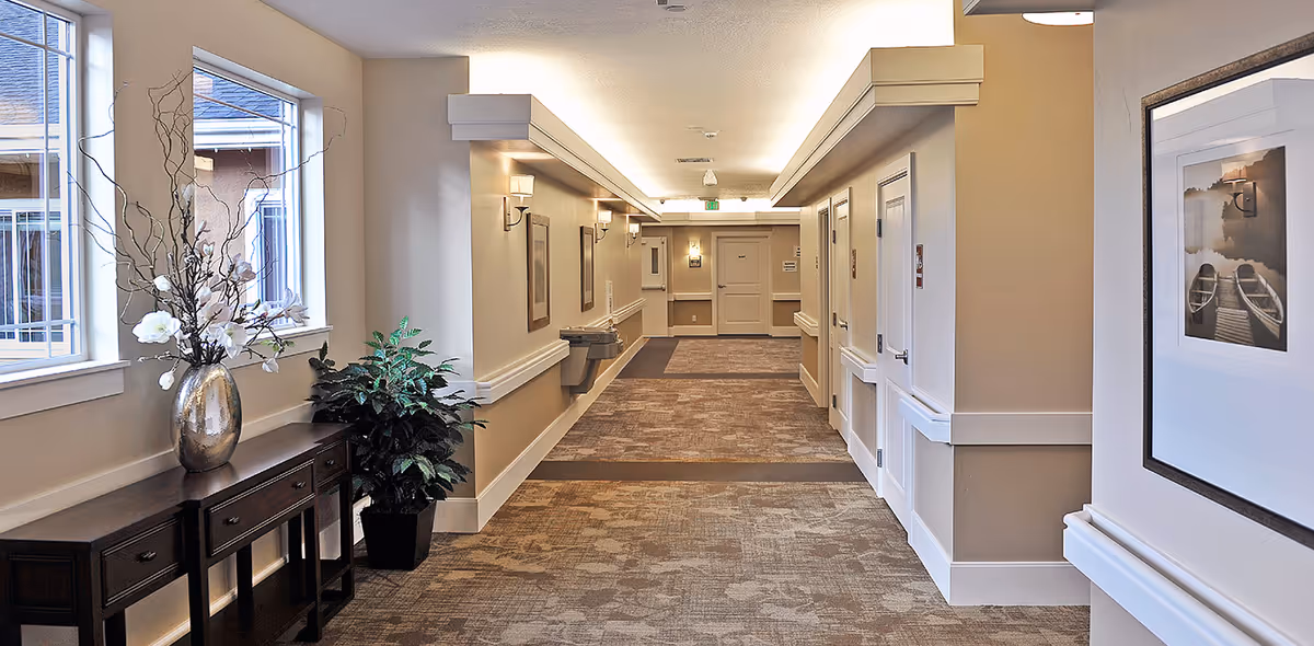 Well-lit carpeted interior hallway with side tables, potted plants, framed artwork, and multiple doors in a senior living facility.