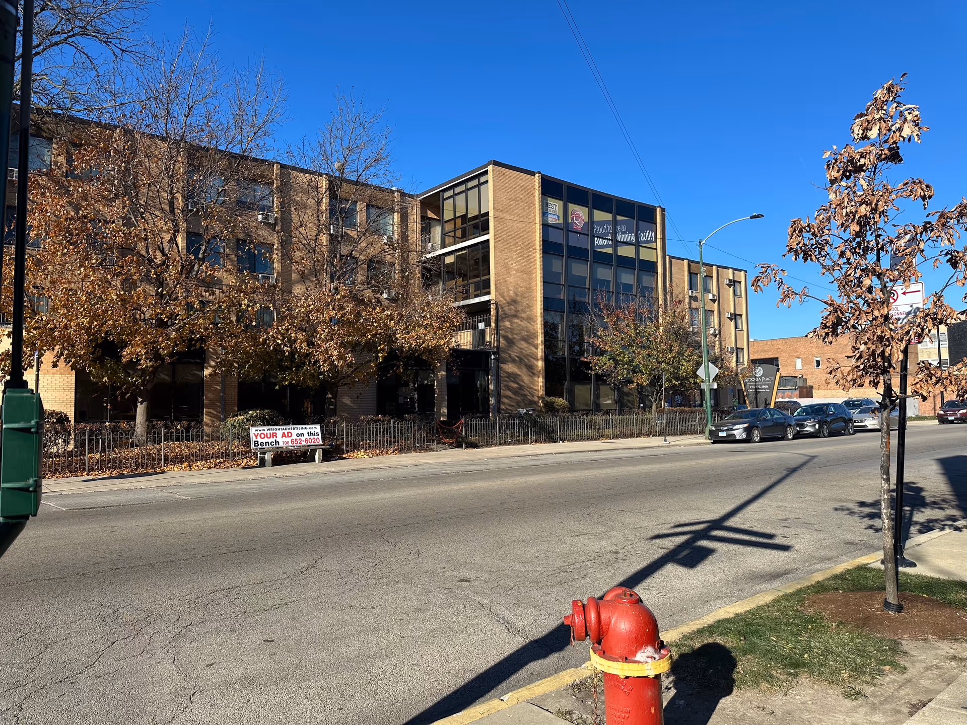 Exterior view of a multi-story brick building with large windows, trees with autumn leaves, parked cars along the street, a red fire hydrant in the foreground, and a clear blue sky.