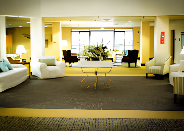 Bright communal lounge with sofas and armchairs arranged around a central table topped with a floral arrangement and large windows in the background.