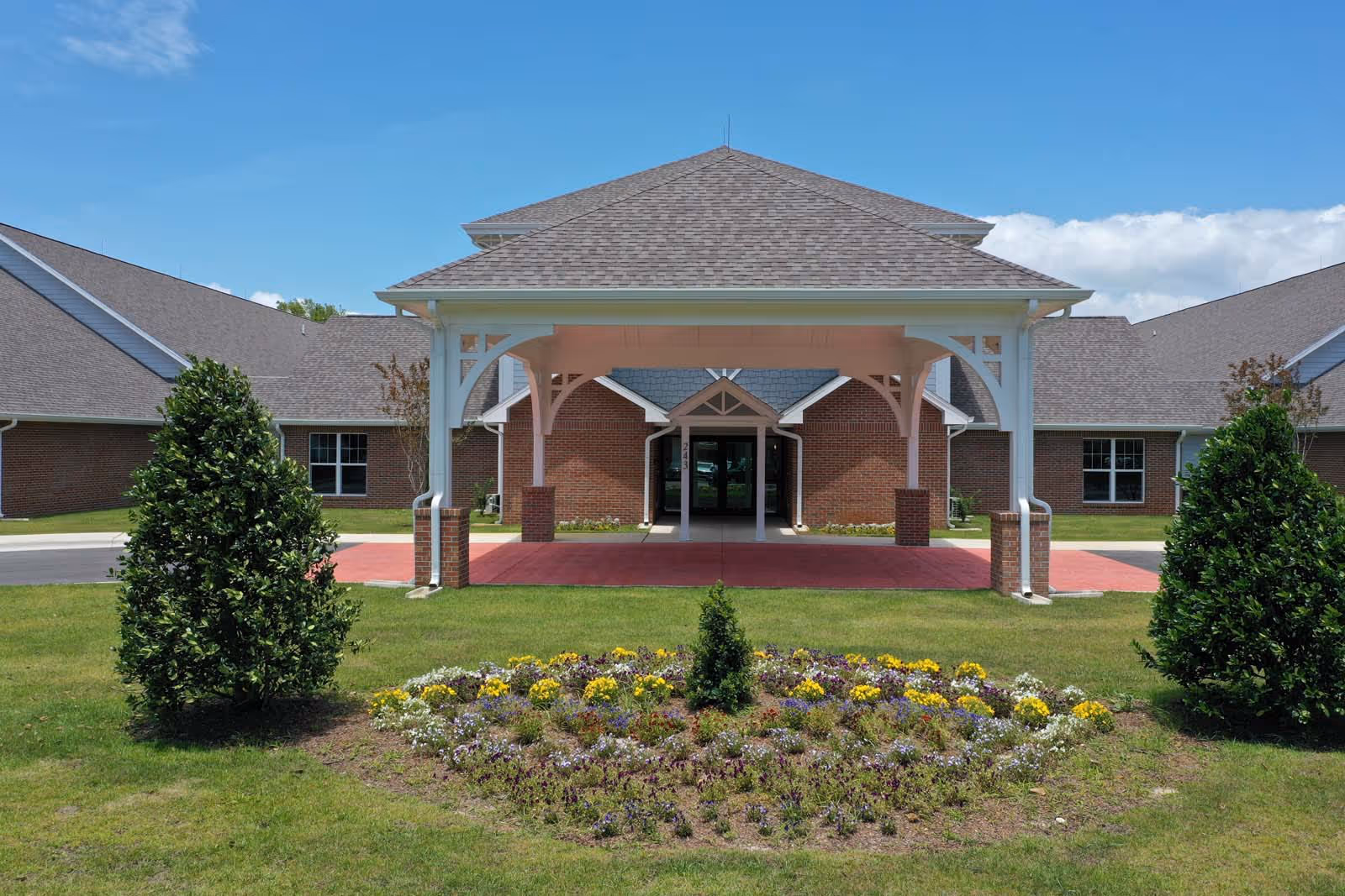 Covered porte-cochere entrance of a brick building with a circular flowerbed and manicured lawn in front.