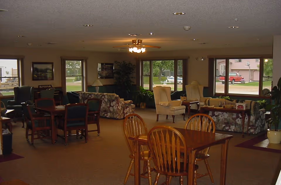 A spacious living room area with multiple seating arrangements including wooden tables with chairs, floral patterned sofas, and beige armchairs. Large windows let in natural light and show a view of the outdoors with parked cars and greenery. The ceiling has recessed lighting and a ceiling fan with lights.