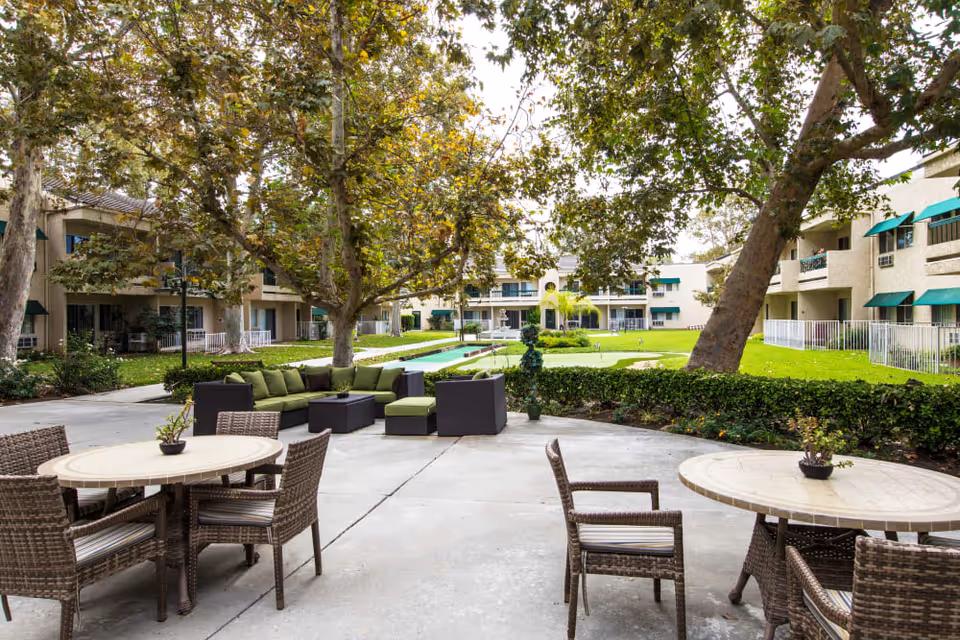 Outdoor patio area at Citrus Place featuring round tables with wicker chairs and a seating area with green cushioned sofas. The patio overlooks a well-maintained lawn with large trees and a fountain in the background, surrounded by a multi-story building with green awnings.