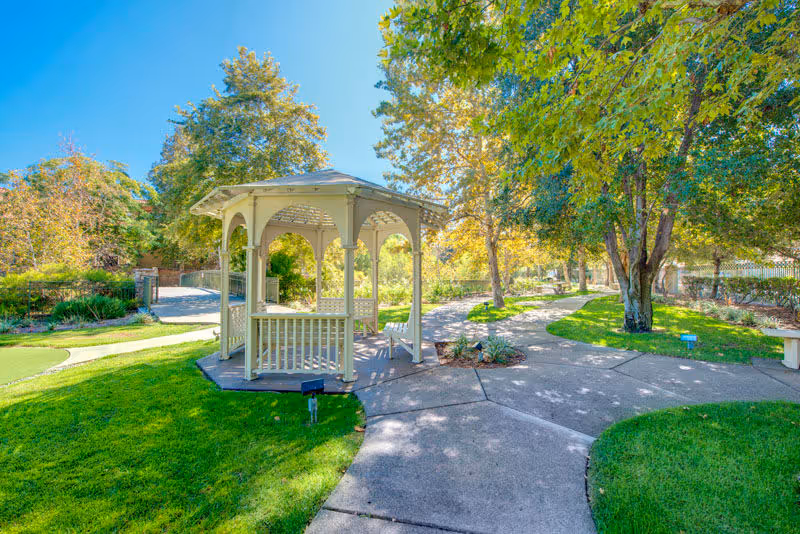 A bright outdoor garden area with a white wooden gazebo surrounded by green grass, trees with autumn foliage, and paved walkways under a clear blue sky.