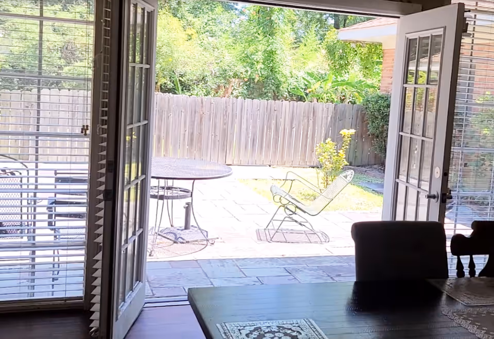 Open French doors from a dining area looking out onto a sunny fenced backyard patio with a metal table and lounge chair.
