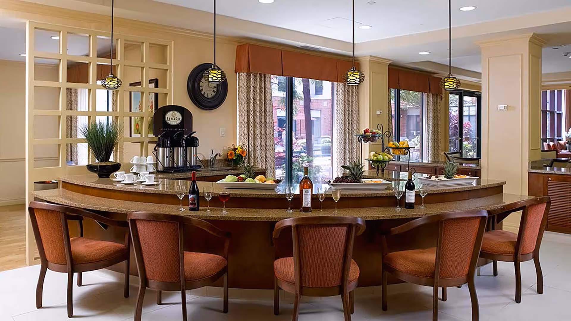 A cozy dining area with a curved granite countertop bar surrounded by six wooden chairs with orange cushions. The countertop has bottles of wine, wine glasses, and plates of fruit. Behind the bar, there is a coffee station with cups and a clock on the wall. Large windows with curtains provide a view of the outdoor garden area.