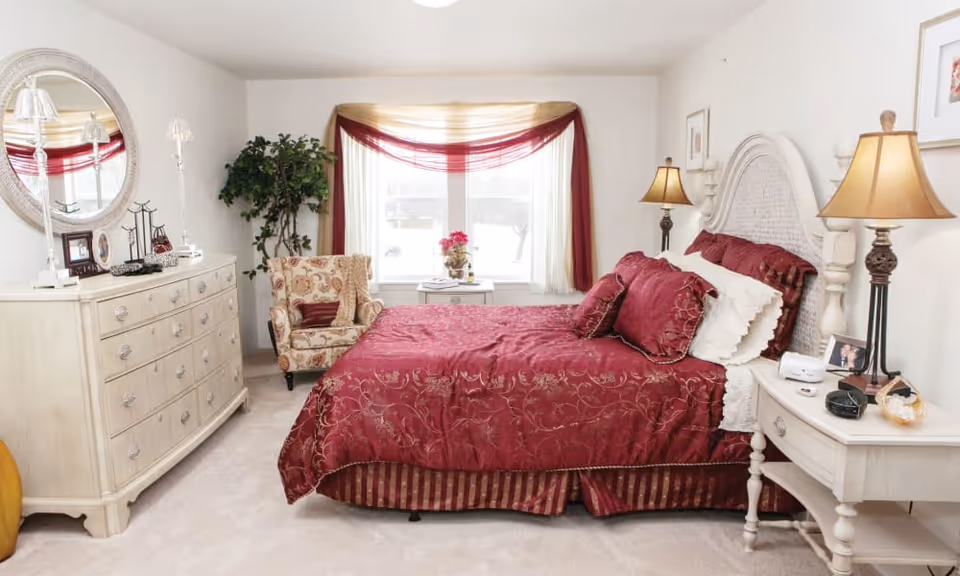 A cozy bedroom with a large bed covered in a red patterned bedspread and matching pillows. The room features a white wooden headboard, two bedside tables with lamps, a white dresser with a round mirror, a patterned armchair, and a window with sheer curtains and red drapes. There is a potted plant in the corner and framed artwork on the walls.