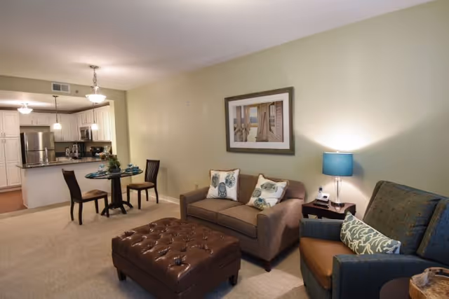 Open-concept living area with a sofa, armchair, tufted ottoman, small dining table, and a kitchen in the background.
