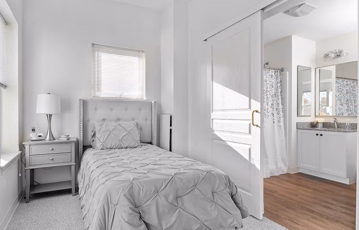 A bright, clean bedroom with a single bed covered in a gray comforter and matching pillow. Next to the bed is a gray nightstand with a lamp, a phone, and a small bowl. A window with blinds is above the bed. To the right, a sliding door is open, revealing a bathroom with a shower curtain, a vanity with a granite countertop, and two mirrors.