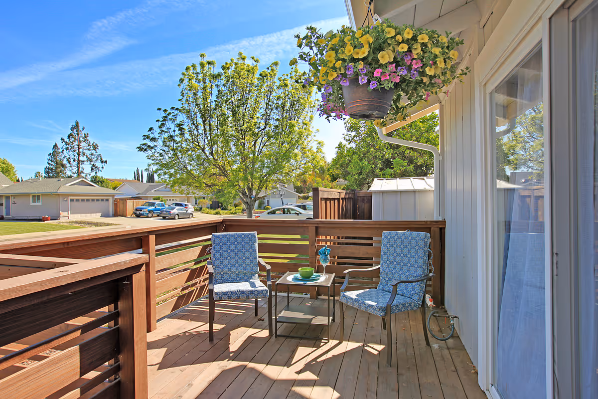 A sunny outdoor wooden deck with two blue cushioned chairs and a small table between them. A hanging flower pot with yellow and purple flowers is attached to the house wall. In the background, there are trees, parked cars, and neighboring houses under a clear blue sky.