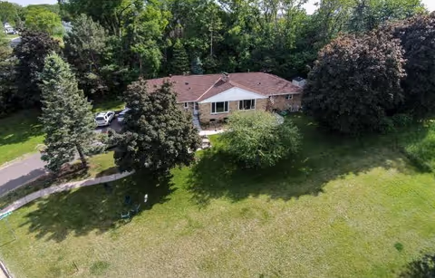 Aerial view of a single-story brick building with a brown roof surrounded by large trees and a well-maintained grassy lawn. There is a paved driveway with a parked white vehicle on the left side of the building.