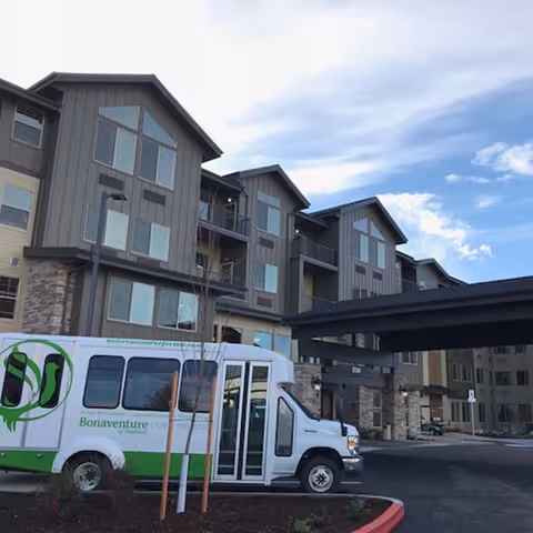 Exterior view of a multi-story senior living facility building with large windows and a covered entrance. A white and green shuttle bus labeled Bonaventure of Medford is parked in front near a landscaped area with small trees. The sky is partly cloudy.