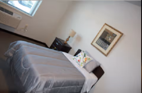 A simple bedroom with a single bed covered in a gray bedspread, a white pillow with a floral design, a bedside table with a lamp, a framed picture on the wall, and a window with an air conditioning unit below it.