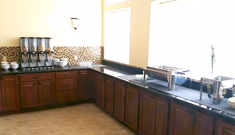 A kitchen area with dark wooden cabinets and black countertops. On the left side, there are three cereal dispensers and several bowls. On the right side, there are two stainless steel food warmers and some plates. The backsplash features small square mosaic tiles in shades of brown and beige. Large windows above the counter let in bright natural light.