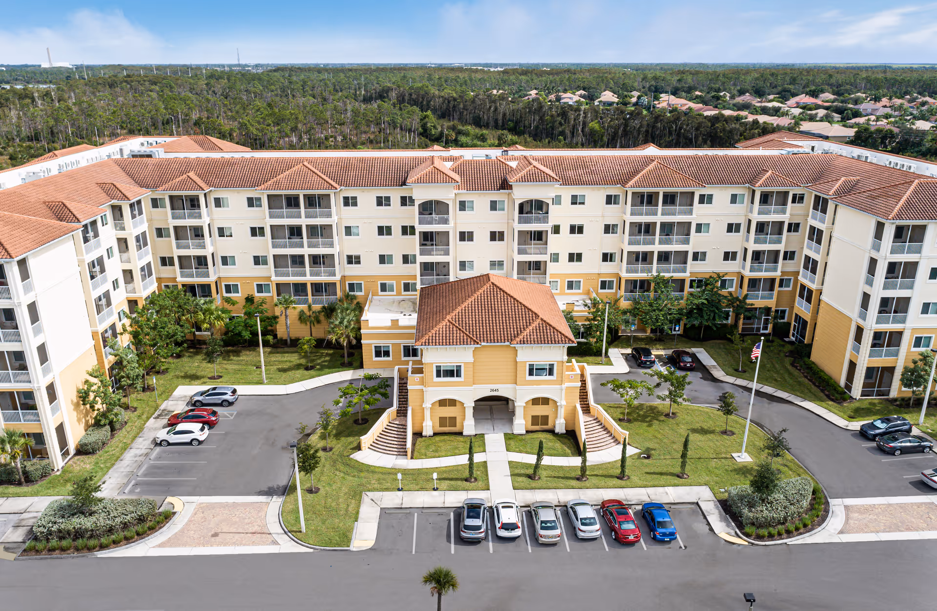 Aerial view of a large senior living facility building with a U-shaped layout, featuring a central entrance with a red-tiled roof and multiple floors with balconies. The building is surrounded by parking spaces, green lawns, and trees, with a wooded area in the background under a partly cloudy sky.