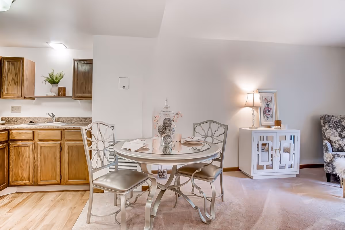A cozy dining area with a round glass-top table set for two, featuring decorative items in the center. Two metal chairs with cushioned seats surround the table. To the left is a kitchen area with wooden cabinets and a countertop with a sink. On the right side, there is a white cabinet with mirrored doors, a table lamp, and a framed floral picture. A floral-patterned armchair is partially visible on the far right.