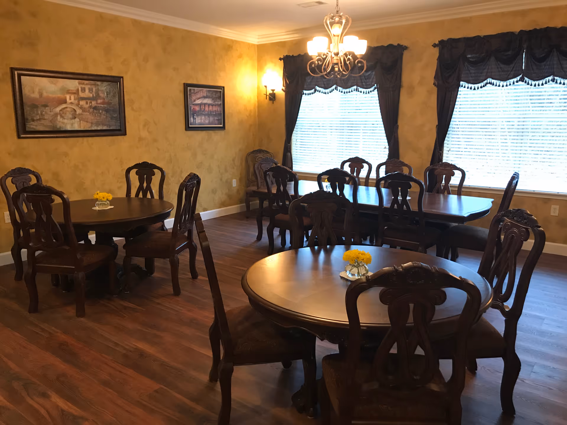 A dining room with multiple wooden tables and chairs arranged neatly. The room has warm yellow walls with two framed paintings, large windows with dark curtains, and a chandelier hanging from the ceiling. Small vases with yellow flowers are placed on the tables.
