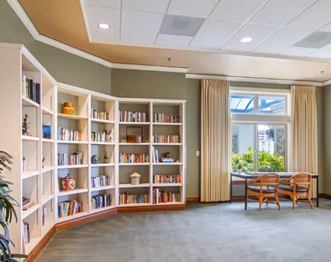 A bright and spacious room with a large built-in bookshelf filled with books and decorative items on the left side. On the right side, there is a window with beige curtains and a small table with two wooden chairs. The room has a light green wall and a carpeted floor.