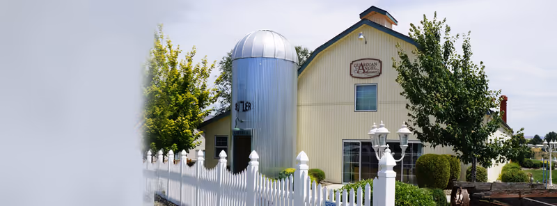 Exterior view of a yellow barn-style building with a silver silo next to it, a white picket fence in front, and green trees and bushes surrounding the area under a partly cloudy sky.