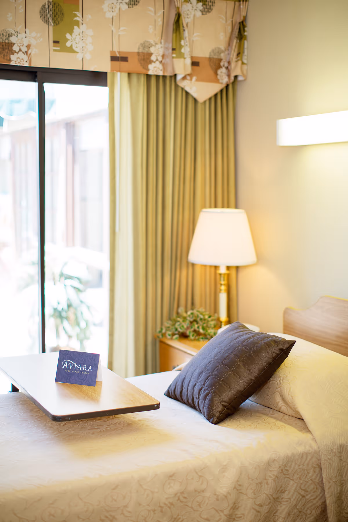 A neatly made bed with a beige patterned bedspread and a dark brown pillow in a well-lit room. A wooden overbed table with a card that reads 'Aviara Healthcare Center' is positioned over the bed. Behind the bed is a nightstand with a lamp and a small plant. The window has green curtains and a floral valance.
