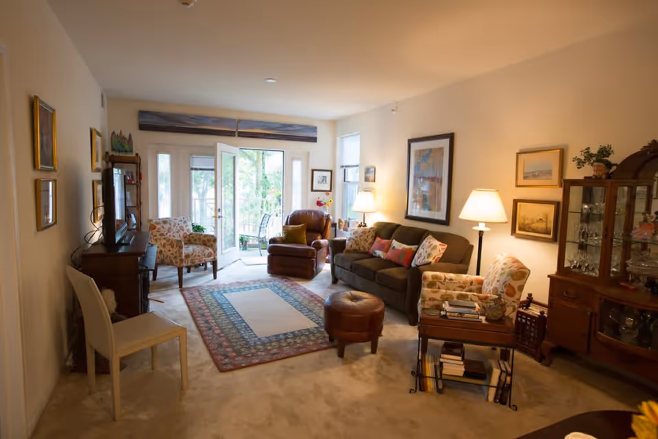 A cozy living room with a brown sofa adorned with colorful pillows, two floral armchairs, a brown recliner, and a round leather ottoman. The room features a patterned area rug on a beige carpet, a wooden cabinet with glassware, framed artwork on the walls, and a TV on a wooden stand. French doors open to a balcony with greenery visible outside.