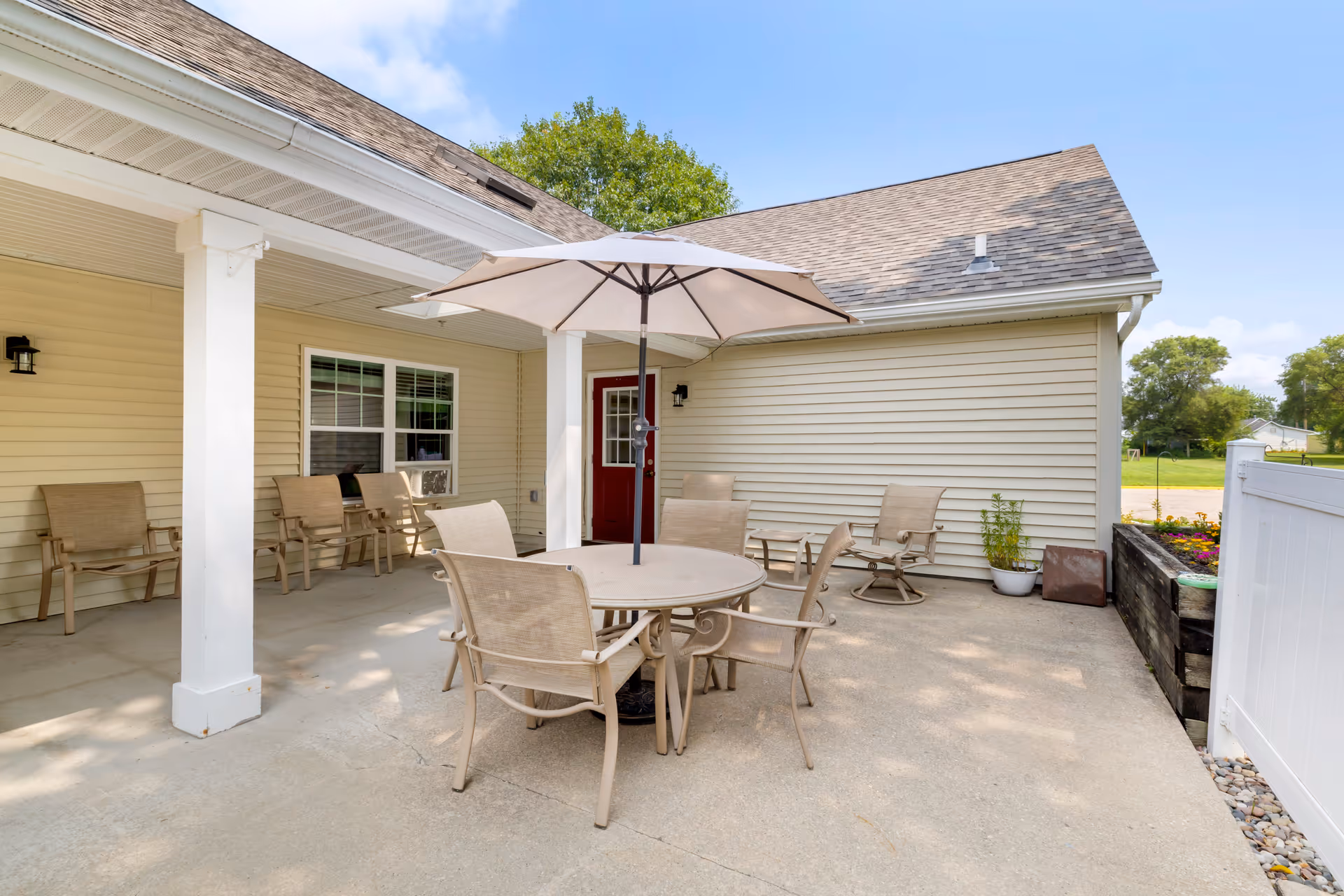 Outdoor covered patio with a round table, umbrella and several chairs beside a beige-sided building with a red door.