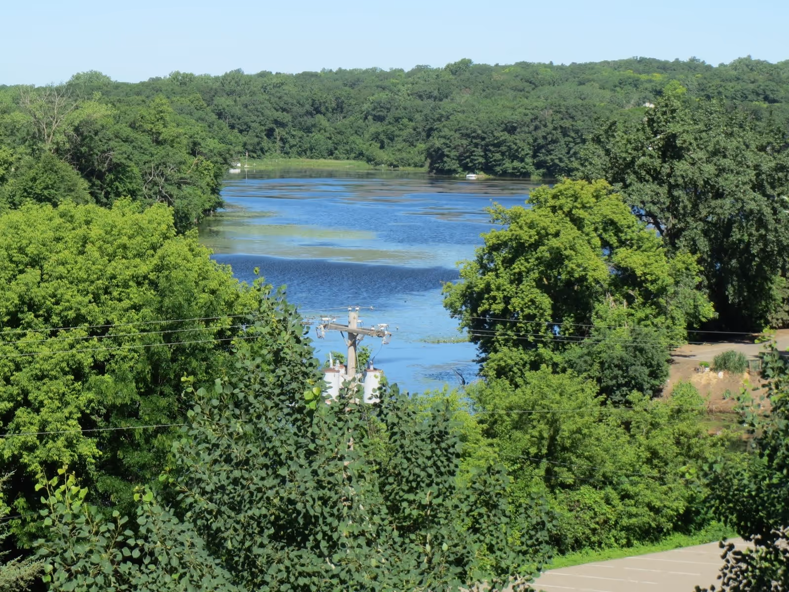 Scenic view of a river surrounded by dense green trees under a clear blue sky, with a utility pole and power lines visible in the foreground.