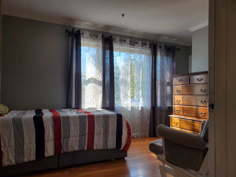 A bedroom with a bed covered in a striped comforter in shades of red, white, black, and gray. There is a wooden dresser with multiple drawers on the right side, and a cushioned chair in front of it. The room has wooden flooring and large windows with sheer and dark curtains allowing natural light to enter.