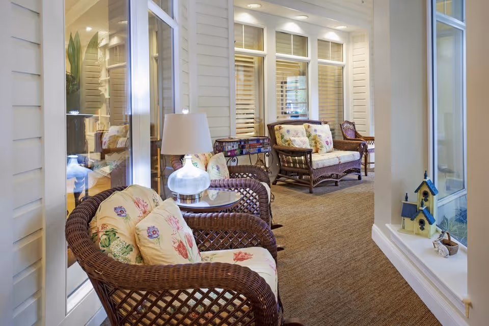 A cozy indoor seating area with wicker chairs and a loveseat, all with floral cushions. A small glass table with a lamp sits between the chairs. The space has large windows with white frames and a beige carpeted floor. Decorative birdhouses are placed on a windowsill to the right.