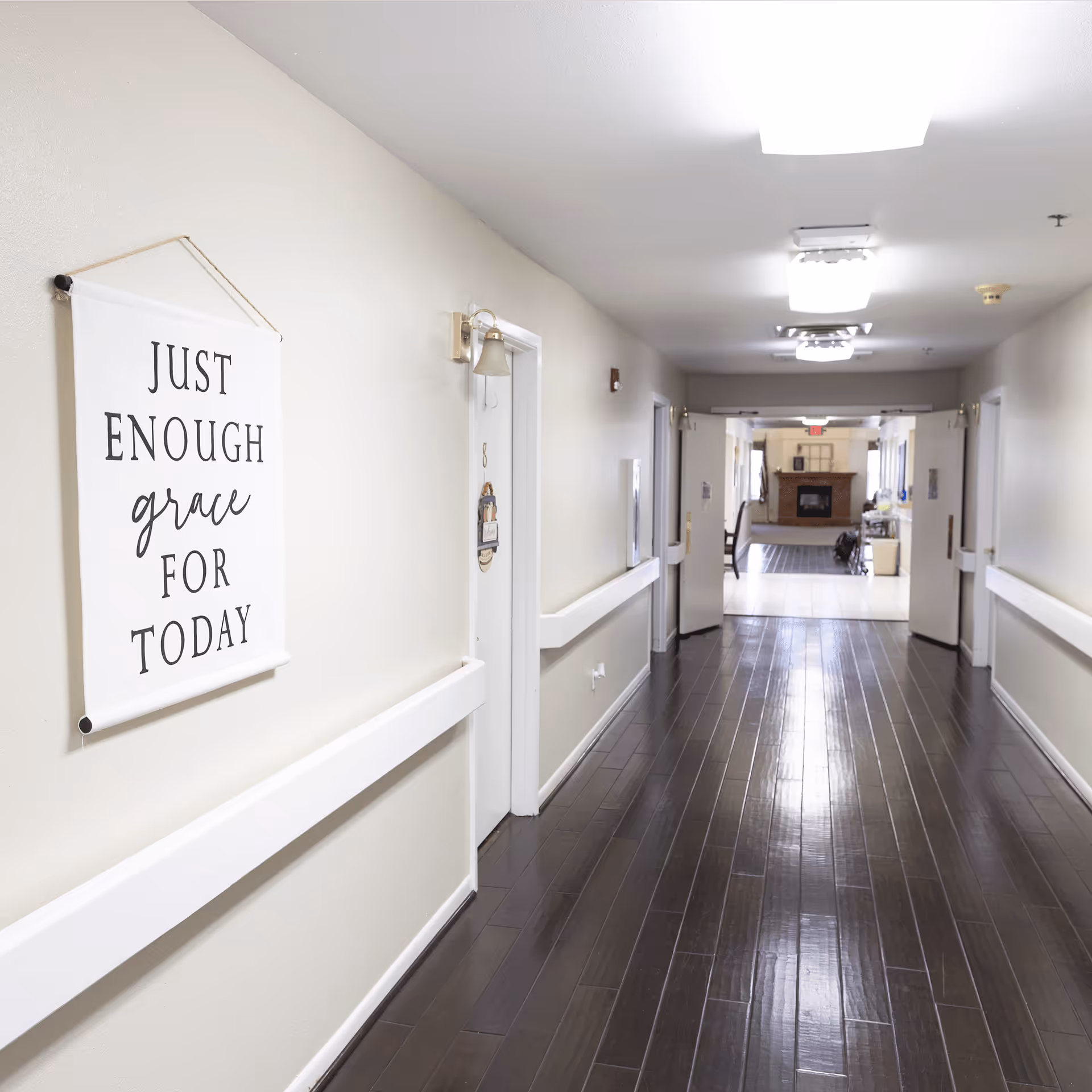 A clean, well-lit hallway in a senior living facility with dark wooden floors and light-colored walls. On the left wall, there is a decorative hanging sign that reads 'JUST ENOUGH grace FOR TODAY'. Several doors line the hallway, and at the end, there is a common area with a fireplace and chairs.
