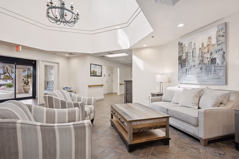 A bright and spacious senior living facility common area with a high ceiling and chandelier. The room features a gray sofa with multiple cushions, two striped armchairs, and a wooden coffee table. A large painting of a cityscape hangs on the wall above the sofa. The floor is tiled, and there is a lamp on a side table next to the sofa. The entrance with glass doors is visible in the background.