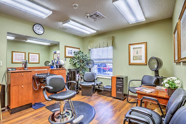 Interior of a hair salon room with wooden flooring, a styling chair in front of a counter with a sink, two black chairs with hair dryers, a window with blinds, framed artwork on light green walls, and a small table with magazines and a flower arrangement.