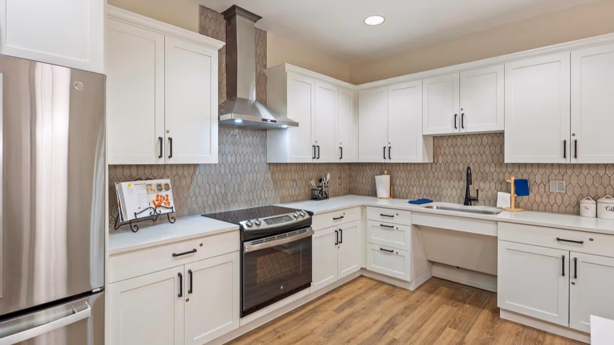 Modern kitchen with white cabinets, stainless steel refrigerator and oven, a range hood, a farmhouse sink, and a backsplash with a geometric tile pattern. The floor is wood, and there is a cookbook stand on the counter.