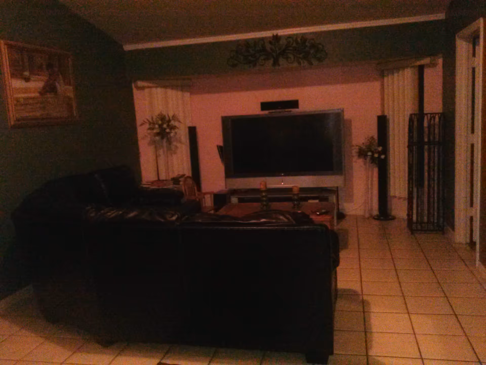 A dimly lit living room with a large flat-screen TV mounted on the wall, a black leather sectional sofa, a coffee table with candles, tiled floor, decorative plants on either side of the TV, and a framed picture on the left wall.