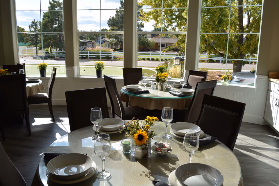 Dining room with round tables set with plates, bowls, wine glasses, and napkins. Each table has a centerpiece with yellow flowers. Large windows in the background show an outdoor view with trees, a white fence, and parked cars.