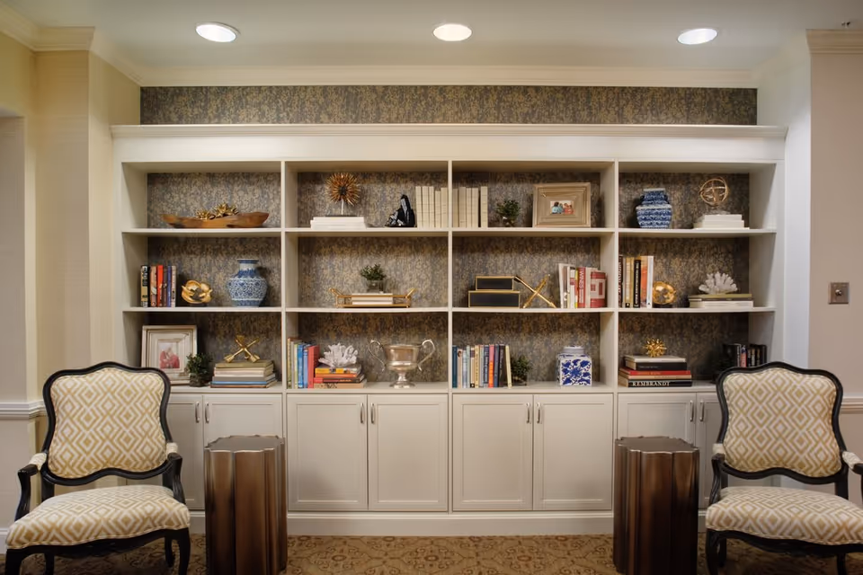 Built-in white shelving unit filled with books and decorative items flanked by two patterned armchairs and small side tables in a sitting area.