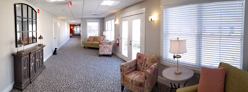 A bright and spacious hallway in an assisted living community with patterned carpet flooring, light-colored walls, and ceiling lights. The hallway features comfortable seating areas with colorful armchairs and sofas, a round side table with a lamp, a large decorative mirror above a wooden cabinet, and large windows with blinds allowing natural light to fill the space. Double glass doors lead outside, and a fire extinguisher is mounted on the wall near the doors.