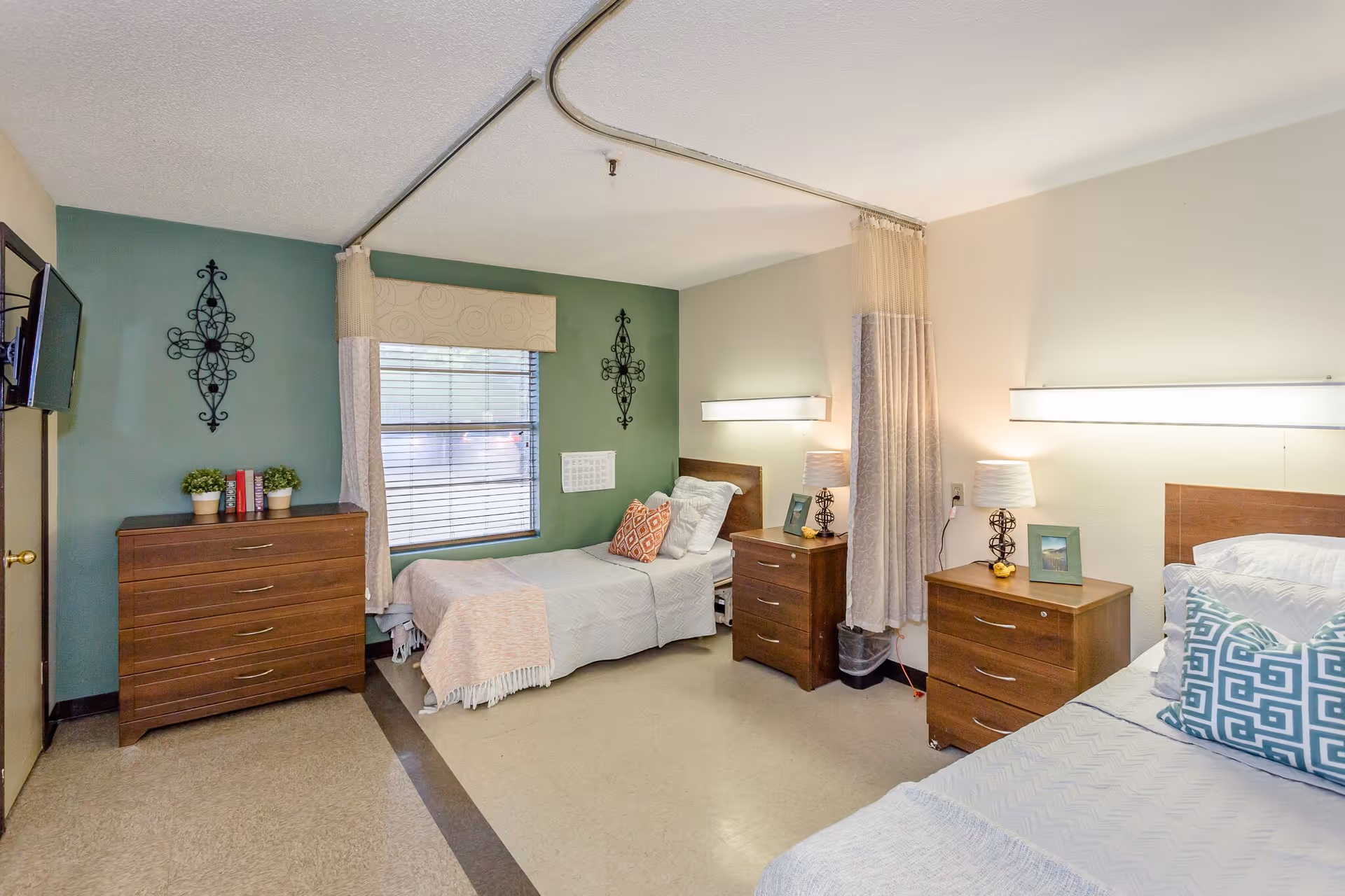 A shared bedroom in a senior living facility with two single beds separated by a privacy curtain. Each bed has a wooden headboard, bedside table with a lamp, and decorative pillows. There is a window with blinds and curtains on the green accent wall, a wooden dresser with potted plants and books, and a wall-mounted TV. The room has a clean and cozy atmosphere.